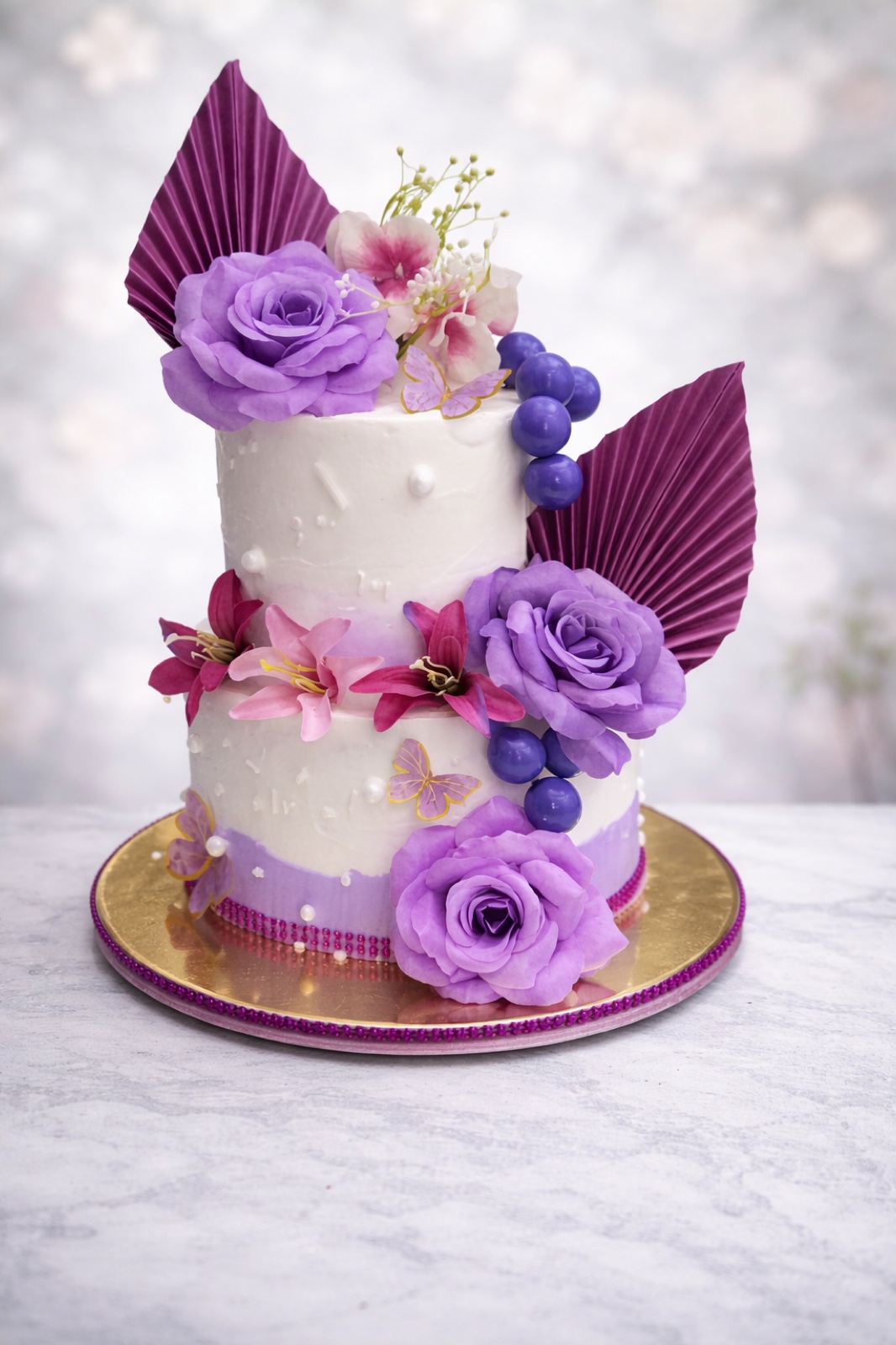 Two-tier white cake with purple sugar roses and gold palm fans